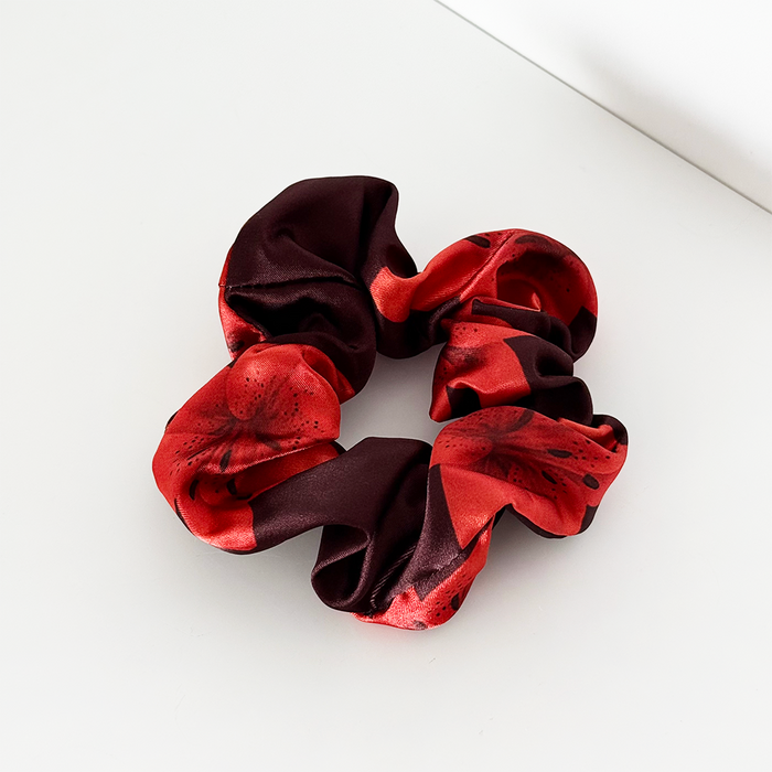 Red and black Coconut Lane Cinnamon Lily Scrunchie on a white surface near the corner.