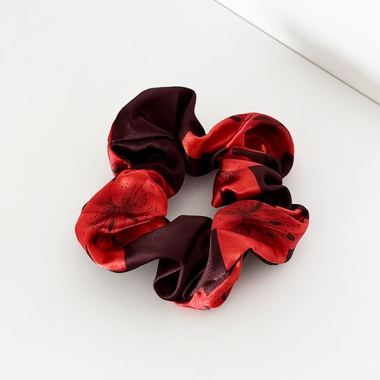 Red and black Coconut Lane Cinnamon Lily Scrunchie on a white surface near the corner.