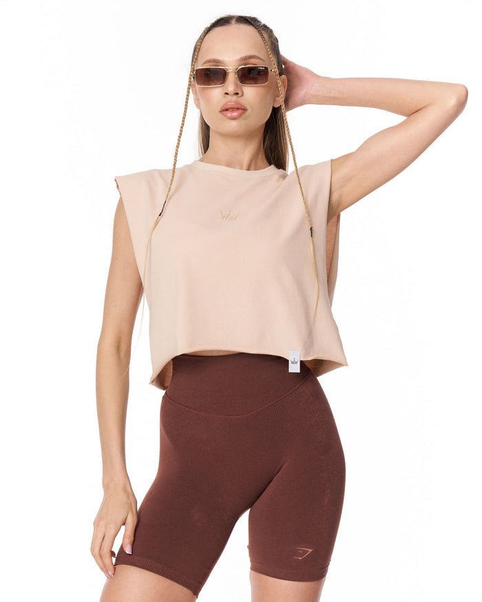 A woman modeling a sleeveless cropped t-shirt in light beige and high-waisted bike shorts in dark brown, styled with sunglasses and a thin gold chain bag against a plain white background.