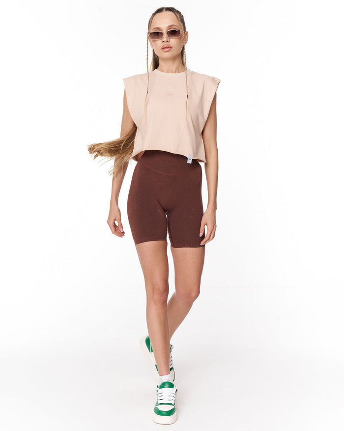 A woman modeling a sleeveless light beige crop top and dark brown bike shorts against a plain white studio background.