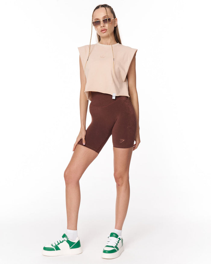 A model wearing a sleeveless cropped top in light beige, dark brown fitted biker shorts, white socks, and green and white sneakers, standing against a plain white background.