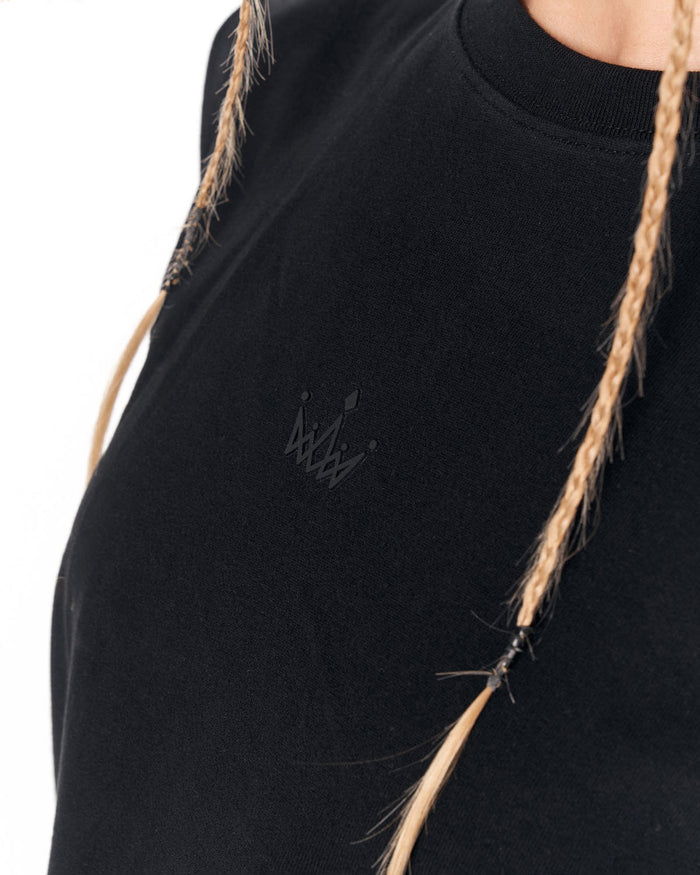 Close-up of a person wearing a black crewneck top with a small tonal embroidered crown logo on the left chest. The person has long blonde braided hair, and the background is solid white.