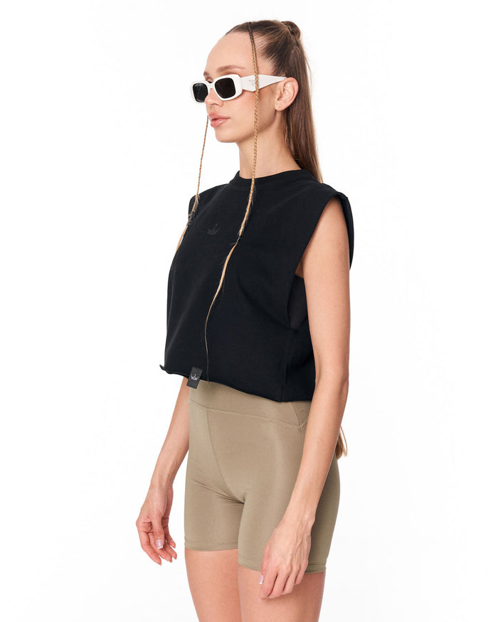 A woman in side profile wearing a black sleeveless cropped zip-front top, beige fitted shorts, and white sunglasses with a chain strap, photographed against a plain white studio background.