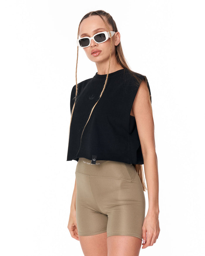 A model wearing a black sleeveless crop top, beige athletic shorts, and white-framed sunglasses with gold chain details, set against a plain white background.