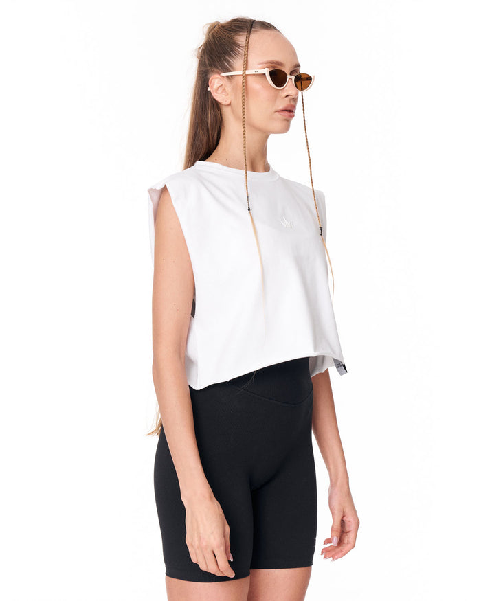 A woman wearing a white sleeveless cropped top, black fitted shorts, and gold-framed sunglasses with thin chains hanging from the temples, photographed against a plain white studio background.