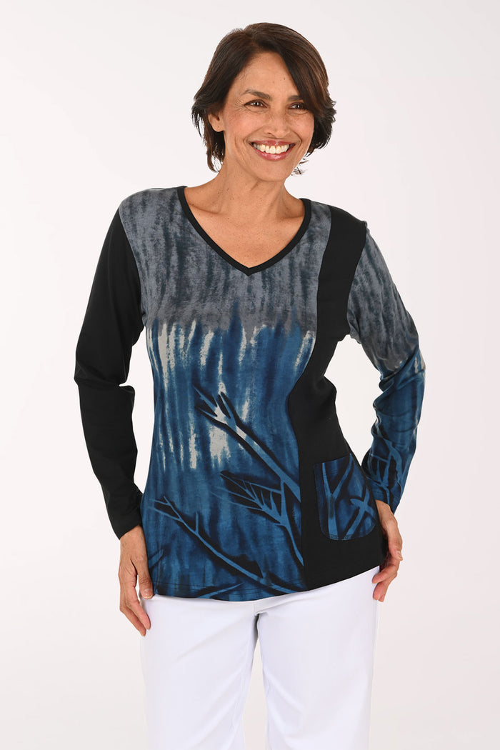 A smiling woman wearing a long-sleeve V-neck top with a blue and black abstract print, paired with white pants, against a plain white background.