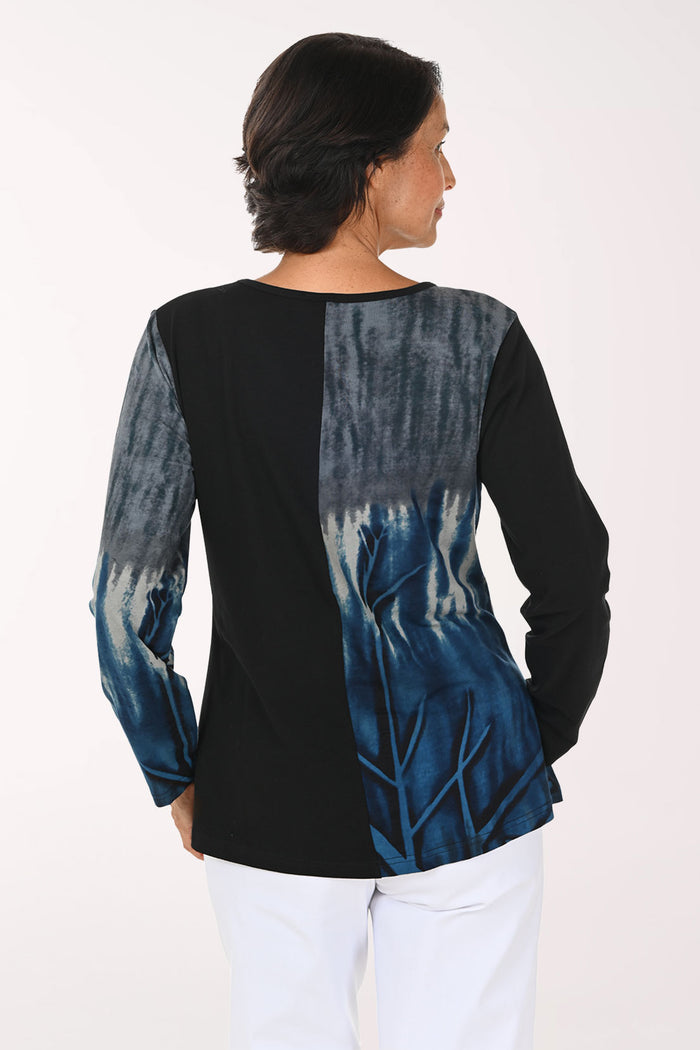 A smiling woman wearing a long-sleeve V-neck top with a blue and black abstract print, paired with white pants, against a plain white background.