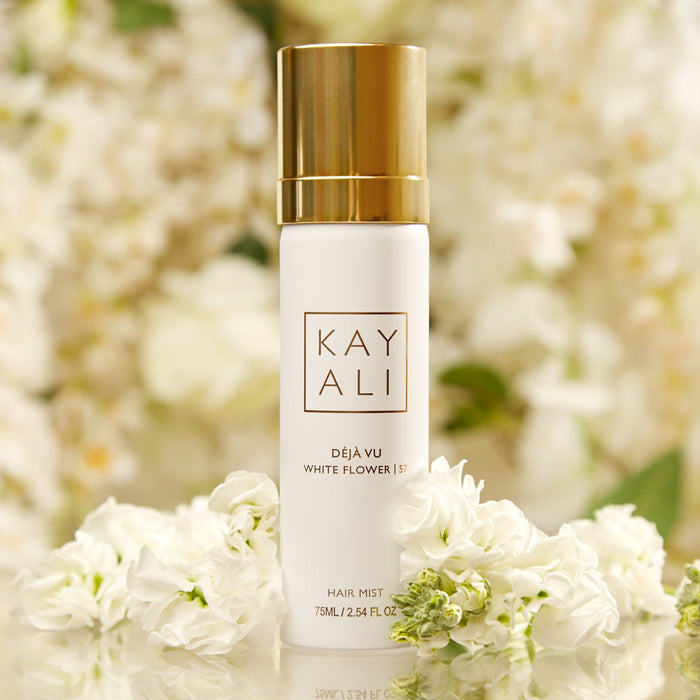 Kayali hair mist bottle with gold cap and white flowers in the background