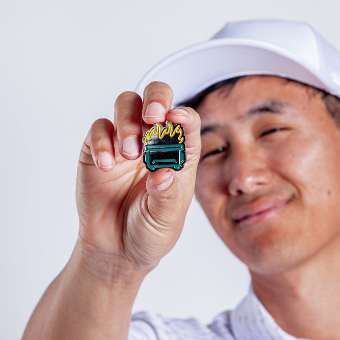 A person in a white cap and shirt smiles while holding the Dumpster Fire - Ball Marker, with their face slightly blurred. The ball marker is in sharp focus, showing green metal and yellow flames with iron nickel plating.