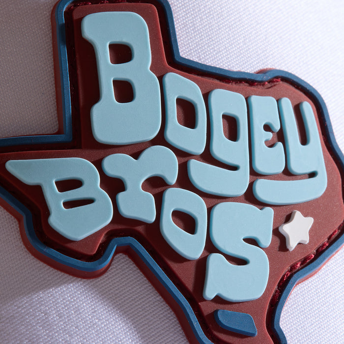 A thick pin shaped like Texas features the words BOGEY BROS in large, light blue letters on a red background, with a small white star under the S. The pin sits on textured white fabric. Total characters: 191.