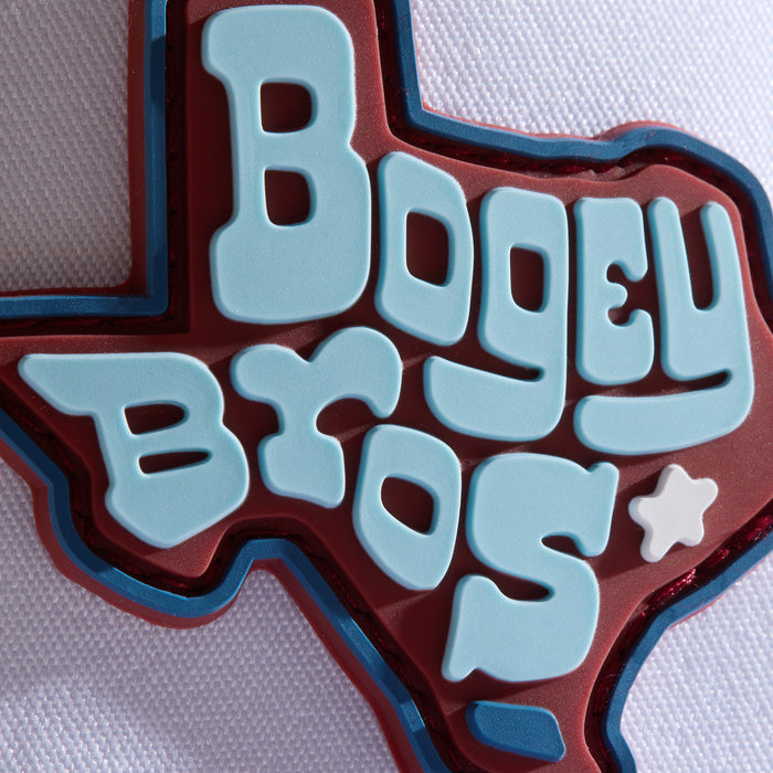 A patch in the shape of Texas features large, light blue bubble letters spelling “BOGEY BROS” on a red background. A white star replaces the “R” in “BROS.” The patch rests on a textured white fabric surface.