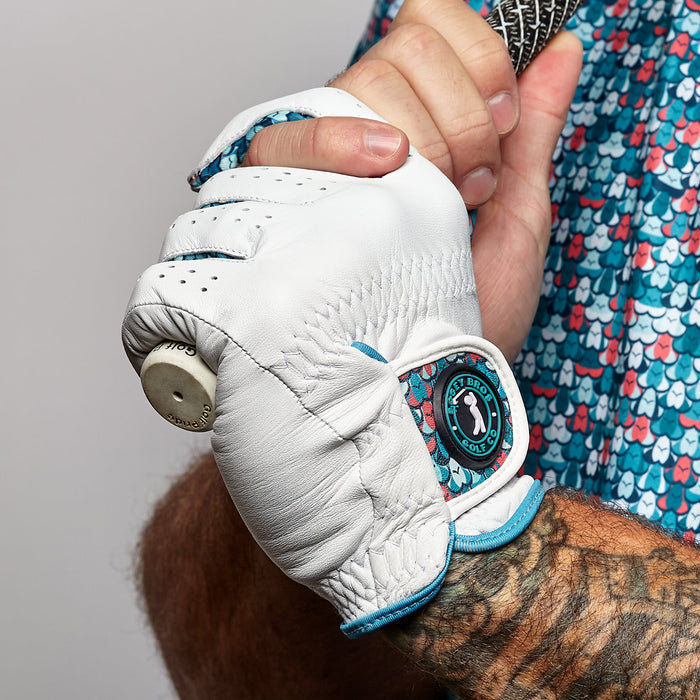 A tattooed arm grips a golf club, wearing the Eyes Up Here - The 305 - Glove, a precision-fit Cabretta leather glove with blue trim and a circular logo. The shirt displays vibrant blue, pink, and red fish scale patterns on a plain background.