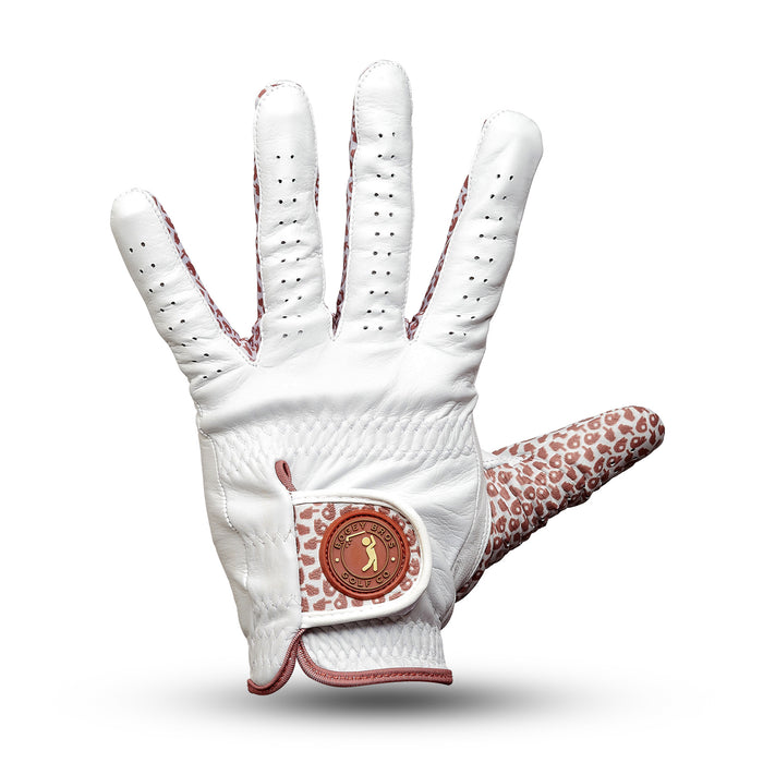 The Get In The Hole - Glove is a white Cabretta leather golf glove with small ventilation holes, featuring pink leopard print accents on the thumb, finger edges, and wrist, plus a circular logo patch and brown stitching details.