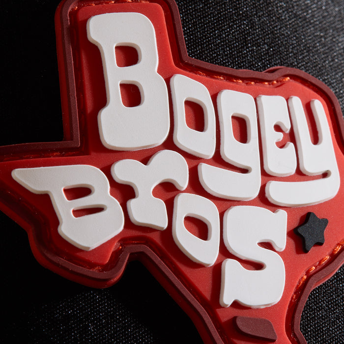 A red Texas-shaped patch with bold, white, playful letters spelling Bogey Bros. A small black star is placed in the lower right area. The patch is sewn onto a textured black fabric background.