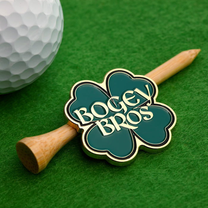 A golf ball sits on green turf near a wooden tee, which holds the Gettin' Lucky - Ball Marker, a four-leaf clover design with gold trim and green fill featuring "BOGEY BROS" in bold white letters across the clover.