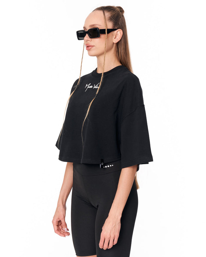 A woman modeling a black cropped zip-up t-shirt with white script embroidery on the chest, paired with black fitted shorts, against a plain white background.
