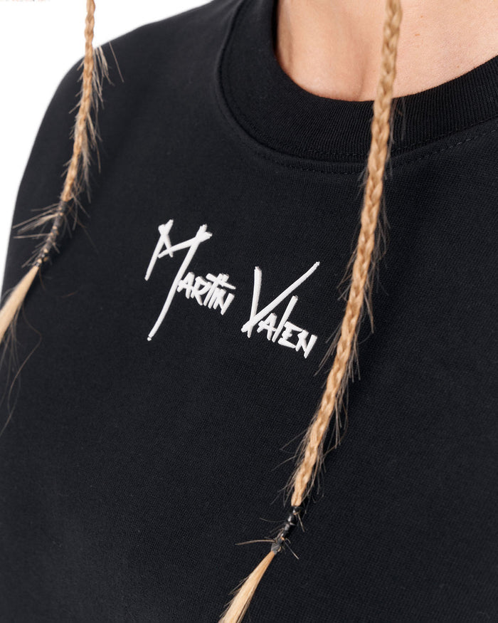 Close-up of a black crewneck sweatshirt featuring a white embroidered 'Martin Valen' logo on the chest, worn by a model with blonde braids.