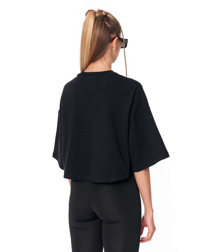 A woman shown from the back wearing a black cropped t-shirt with wide, short sleeves and black leggings, photographed against a solid white background.