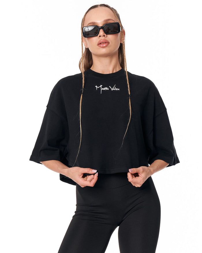 A model wearing a black cropped t-shirt with a white script logo on the chest, paired with black leggings and black sunglasses, against a plain white background.