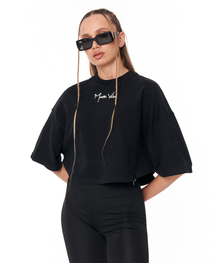 A model wearing a black cropped t-shirt with a white script logo on the chest, paired with black leggings and black sunglasses, set against a plain white background.
