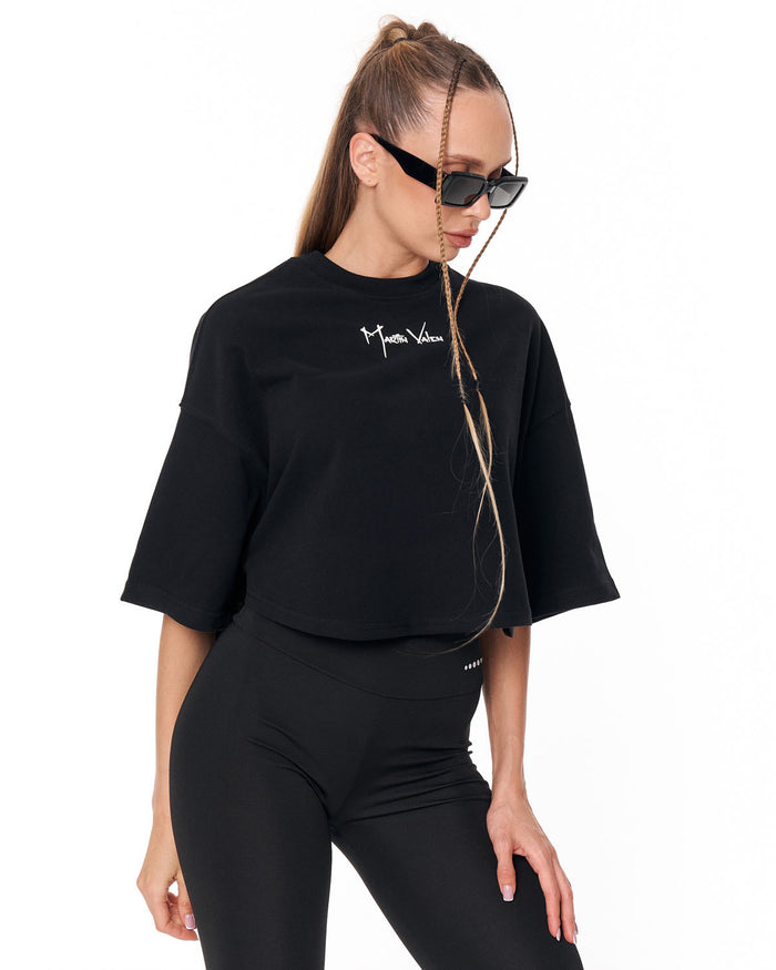 A woman modeling a black cropped t-shirt with a white script logo on the chest, paired with black leggings, against a plain white background.