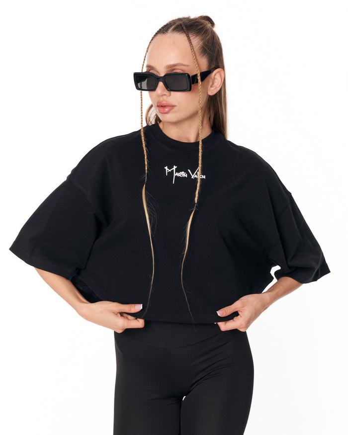 A woman modeling a black oversized short-sleeve top with a white 'Martin Valen' logo on the chest, paired with black pants, against a plain white background.