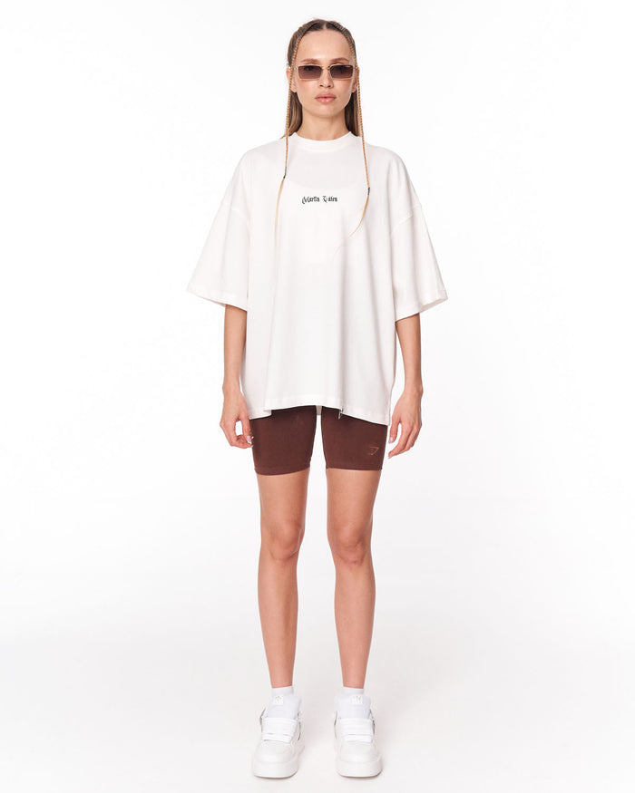 A model stands against a white studio background wearing an oversized white t-shirt with 'johnnie life' printed in black on the chest, brown fitted shorts, white sneakers, and sunglasses.
