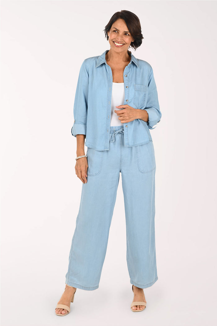A woman modeling a light blue denim wide-leg pant, styled over a white tank top and beige sandals against a plain white studio background.