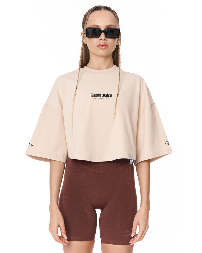 A model wearing a light beige cropped t-shirt with 'Martin Valen' printed on the chest, paired with dark brown biker shorts, against a plain white background.