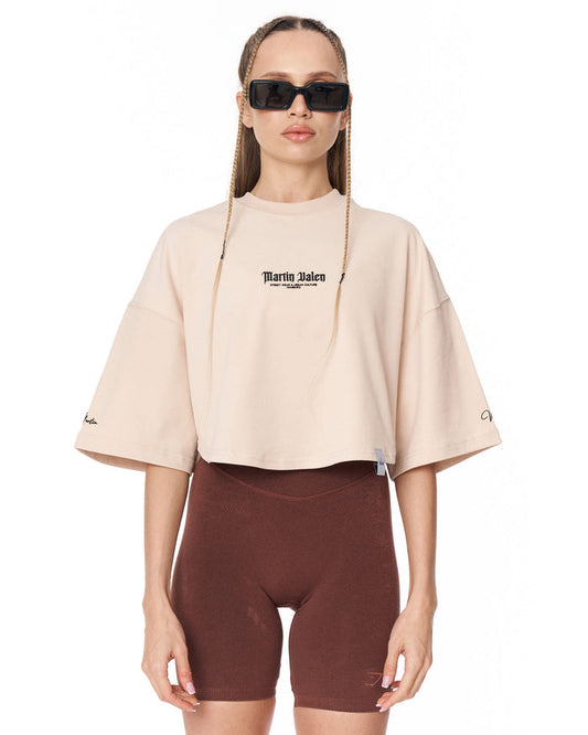 A model wearing a light beige cropped t-shirt with 'Martin Valen' printed on the chest, paired with dark brown biker shorts, against a plain white background.