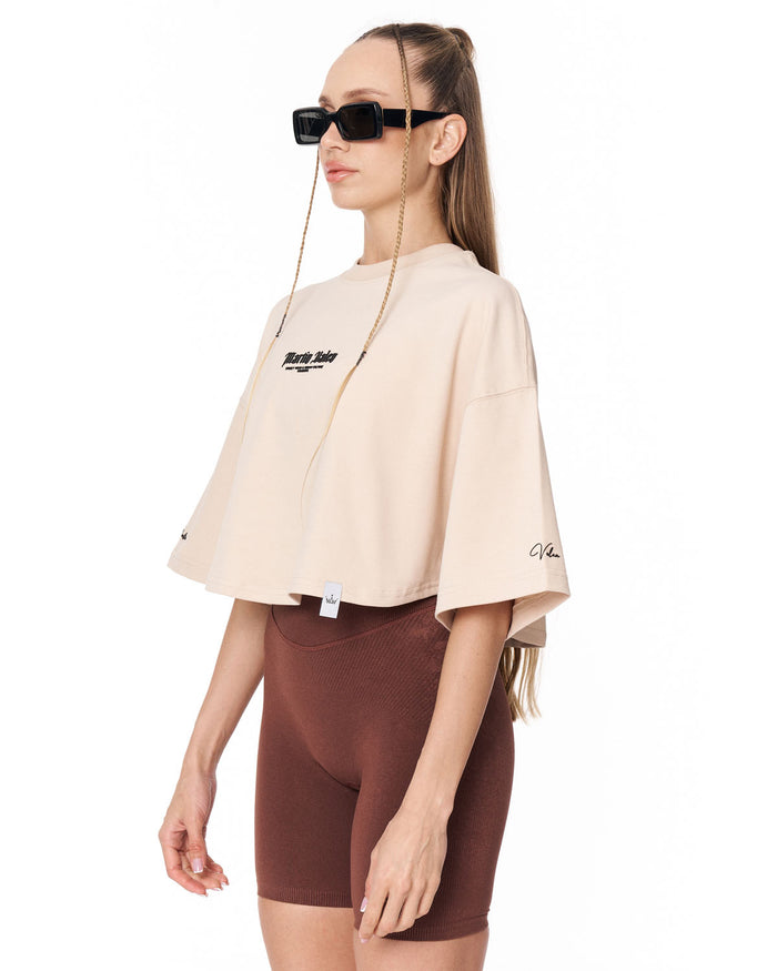 A woman modeling a beige cropped t-shirt with black graphic text on the chest, paired with brown fitted shorts, against a plain white background.