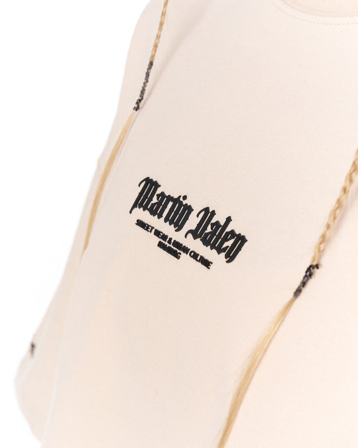 Close-up of a light cream or off-white garment with black gothic-style 'Martin Valen' text printed on the chest, with thin gold-toned braided cords hanging near the shoulder area.