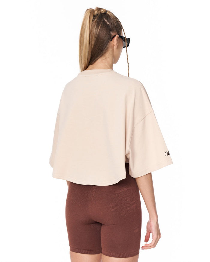 A woman shown from the back wearing a light beige cropped t-shirt with wide three-quarter sleeves and brown fitted bike shorts, against a plain white background.