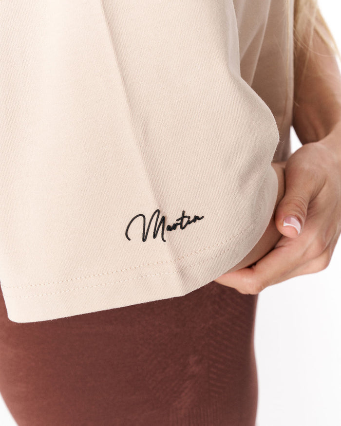 Close-up of a person wearing a light beige ribbed knit top with a black script logo reading 'Martin Valen' on the lower left side, paired with brown ribbed bottoms against a white background.