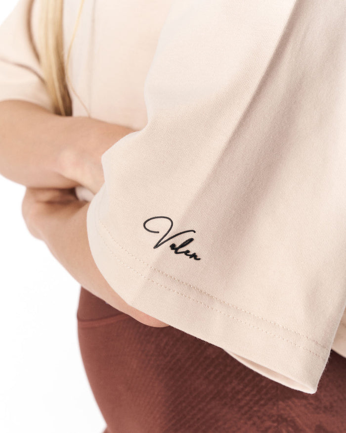 Close-up of a person wearing a cream-colored t-shirt with a black cursive 'Valen' logo embroidered on the sleeve, paired with brown pants.