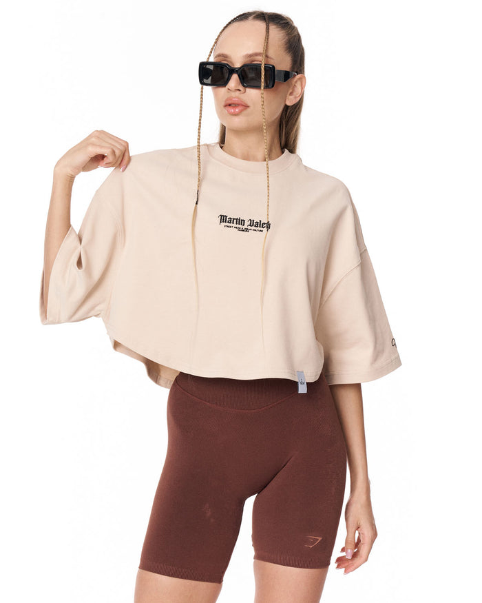 A model wearing a cropped beige t-shirt with 'Martin Valen' printed on the chest, paired with brown bike shorts and black sunglasses, against a plain white background.