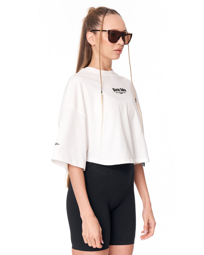 A model wearing a white cropped t-shirt with a black 'Martin Valen' logo on the chest, paired with black bike shorts and sunglasses with chain straps, against a plain white background.
