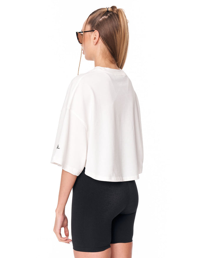 A woman shown from the back wearing an oversized white short-sleeve crop top and black fitted biker shorts against a plain white studio background.