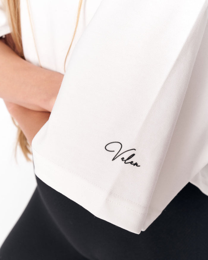 Close-up of a white short-sleeve top worn by a model, featuring a black cursive 'Valen' script logo embroidered on the sleeve cuff, paired with a black bottom.