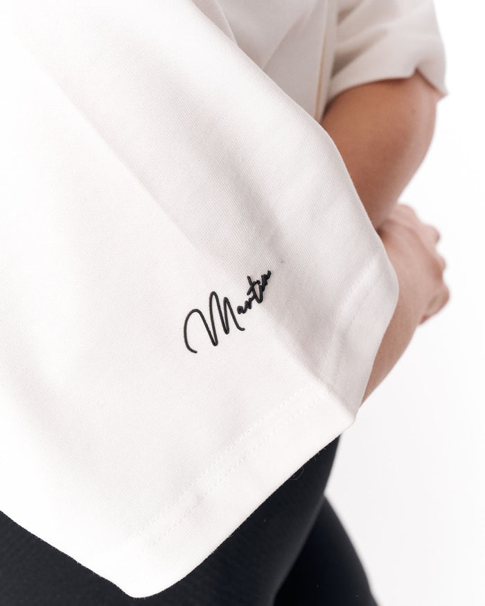Close-up of a person wearing a white short-sleeve top with a black cursive signature-style logo on the sleeve, paired with black bottoms, against a white background.