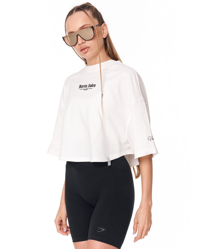 A woman modeling a white cropped zip-up hoodie with 'Martin Valen' printed on the chest, paired with black bike shorts against a plain white background.