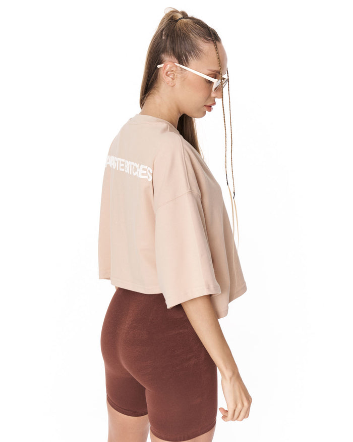 A woman modeling a sleeveless cropped t-shirt in light beige and high-waisted bike shorts in dark brown, styled with sunglasses and a thin gold chain bag against a plain white background.