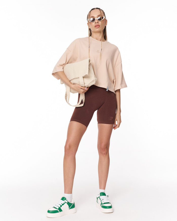A woman modeling a beige crop top, brown fitted biker shorts, white socks, and green and white sneakers, photographed against a plain white studio background.