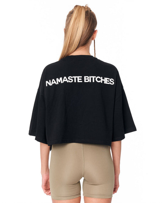 A woman shown from the back wearing a black cropped T-shirt with the phrase 'NAMASTE' printed in bold white letters across the upper back, paired with beige fitted shorts against a plain white background.