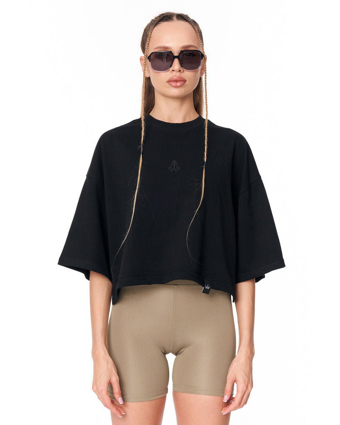 A model wearing a black cropped t-shirt with wide short sleeves and beige fitted biker shorts, styled with black sunglasses against a plain white background.