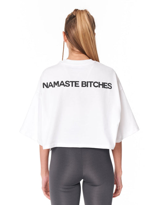 A woman viewed from the back wearing a white cropped T-shirt with the text 'NAMASTE' printed in black across the upper back, paired with dark gray leggings, against a plain white background.