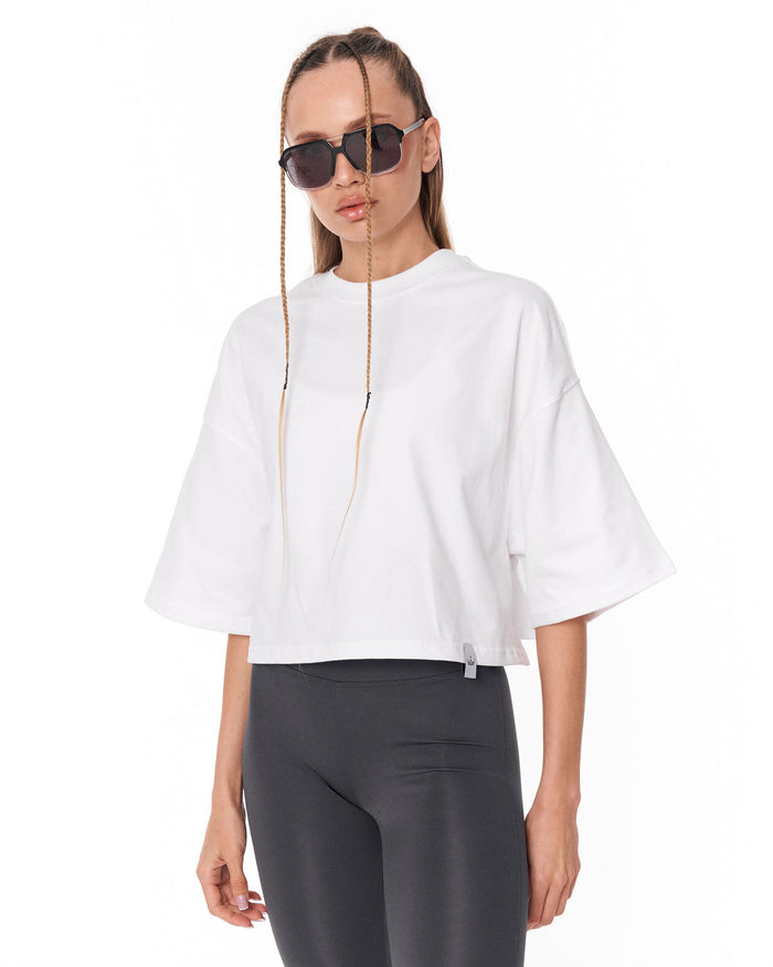 A woman modeling a white cropped t-shirt with wide three-quarter sleeves and dark gray leggings, wearing sunglasses with long hanging chains, against a plain white background.