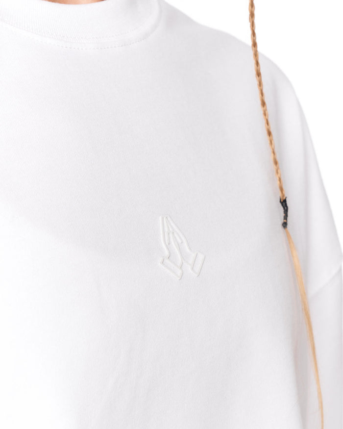 Close-up of a white crewneck sweatshirt featuring a tonal embroidered praying hands logo on the left chest, with a braided tan strap running vertically along the right side.