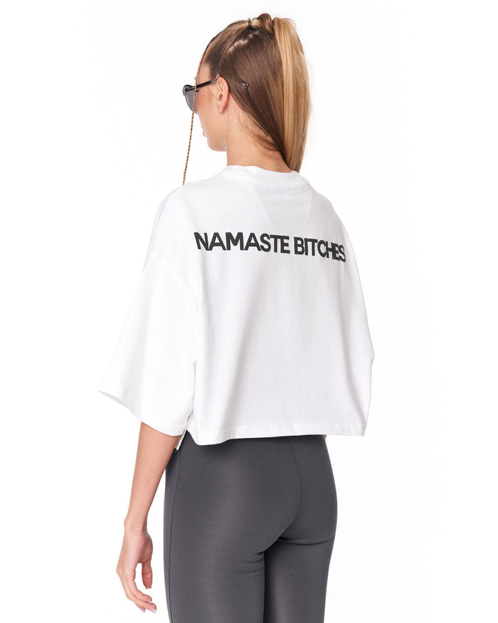 A woman shown from the back wearing a white cropped t-shirt with the text 'NAMASTE.' printed in black across the upper back, paired with dark gray leggings against a plain white background.