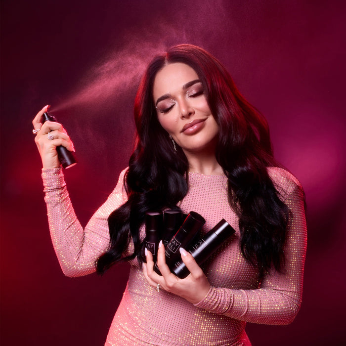 Mona Kattan holding a hair spray bottle and a hair product container against a dark background
